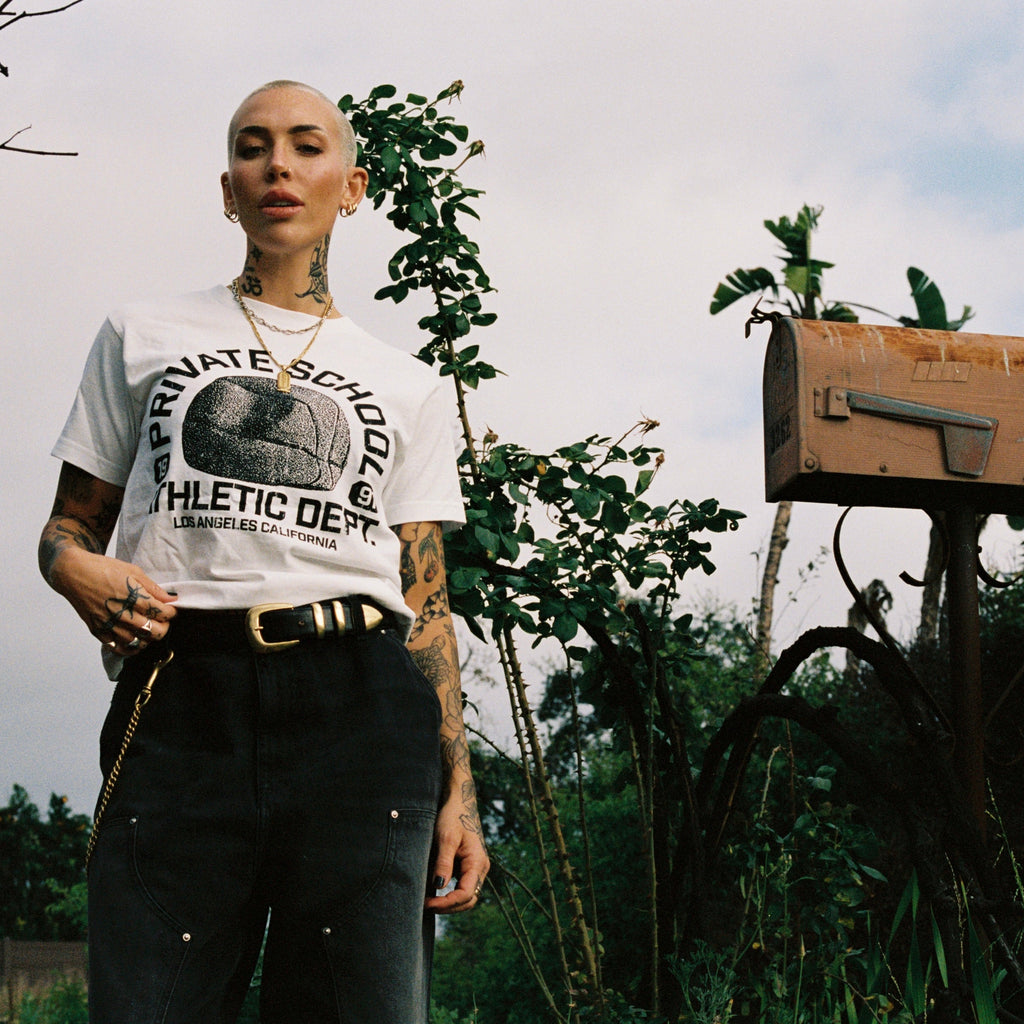 Person wearing a white t-shirt with text and graphics, standing outdoors with greenery and a mailbox in the background.