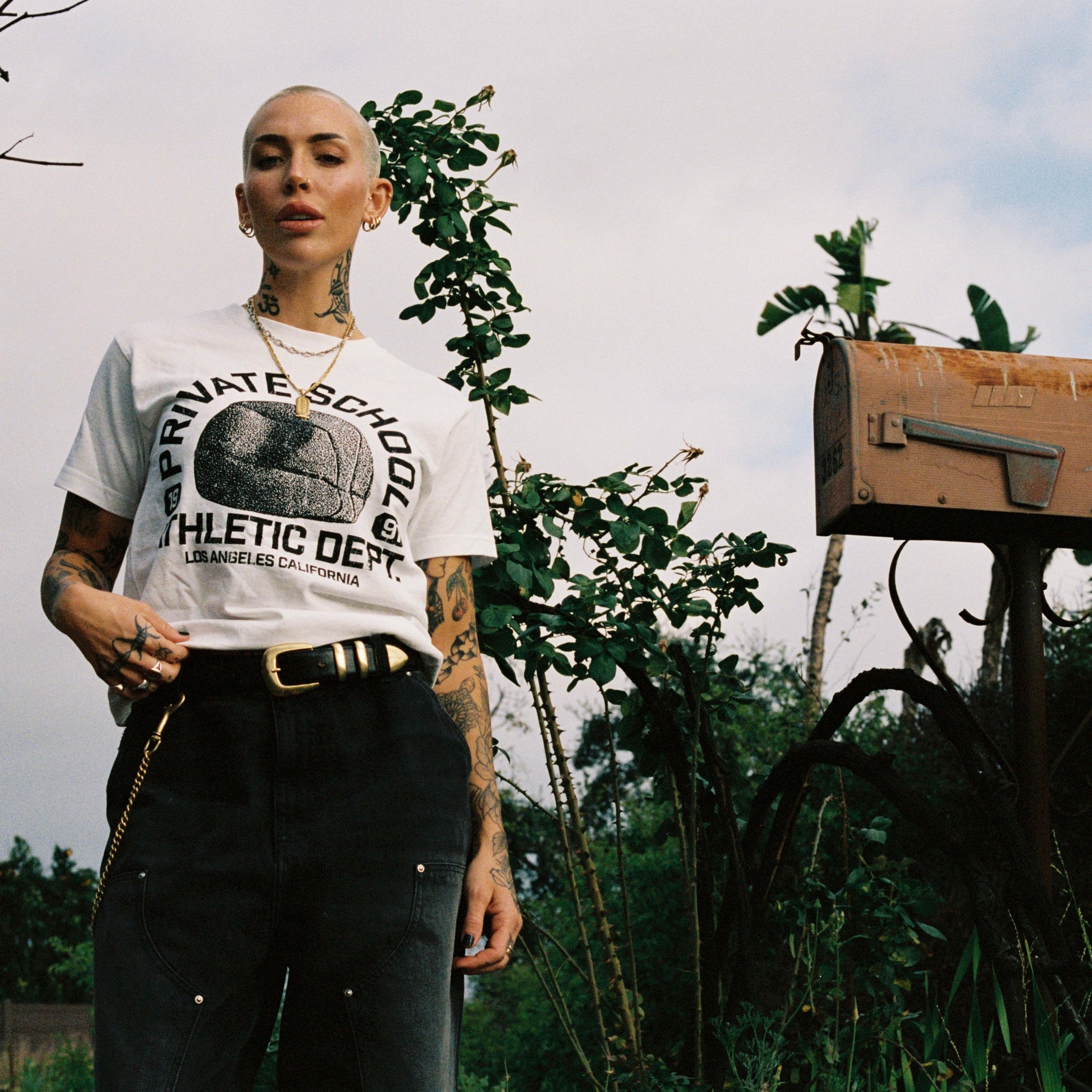 Person wearing a white t-shirt with text and graphics, standing outdoors with greenery and a mailbox in the background.