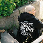 Person wearing a black hoodie with text sitting on a stone ledge outdoors.
