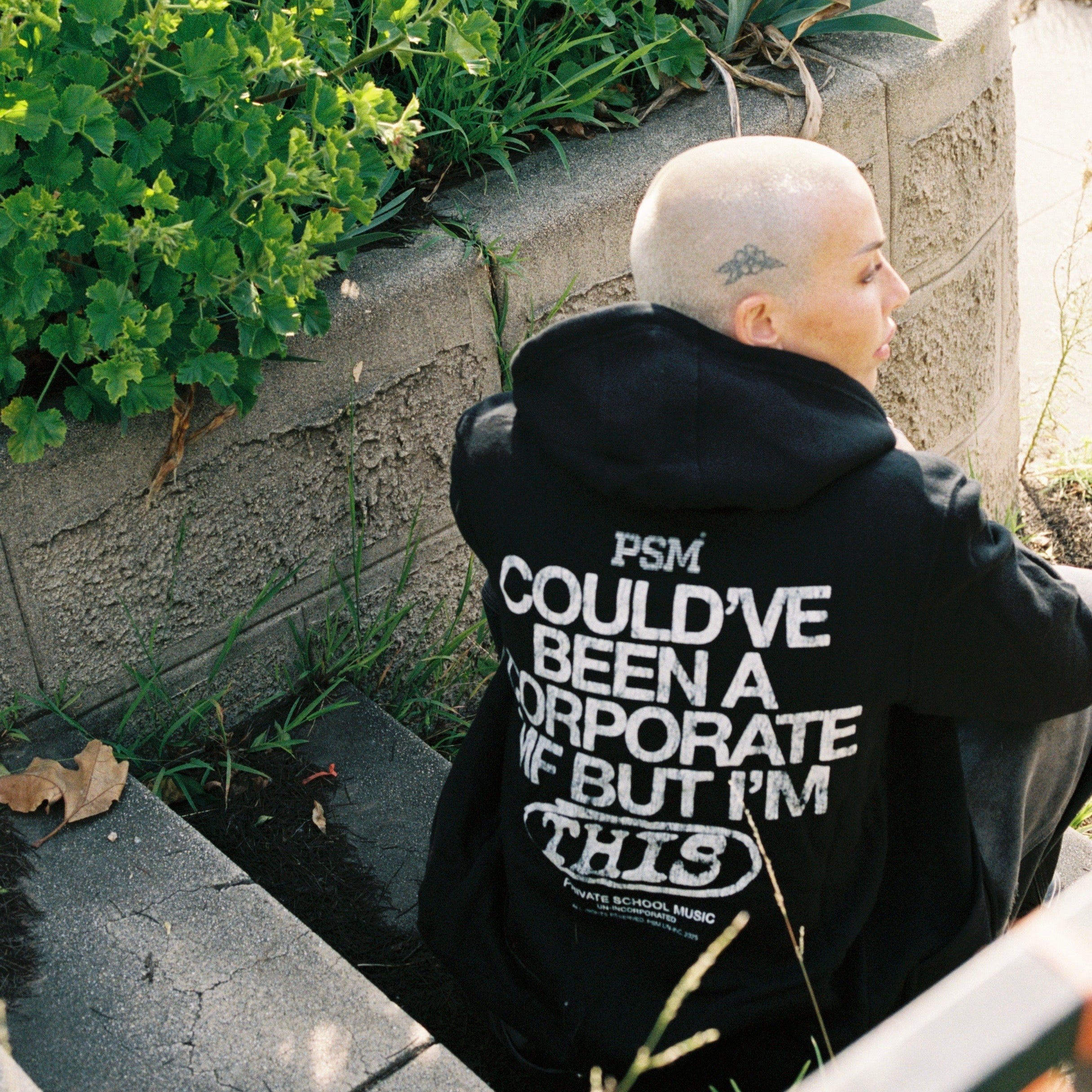Person wearing a black hoodie with text sitting on a stone ledge outdoors.